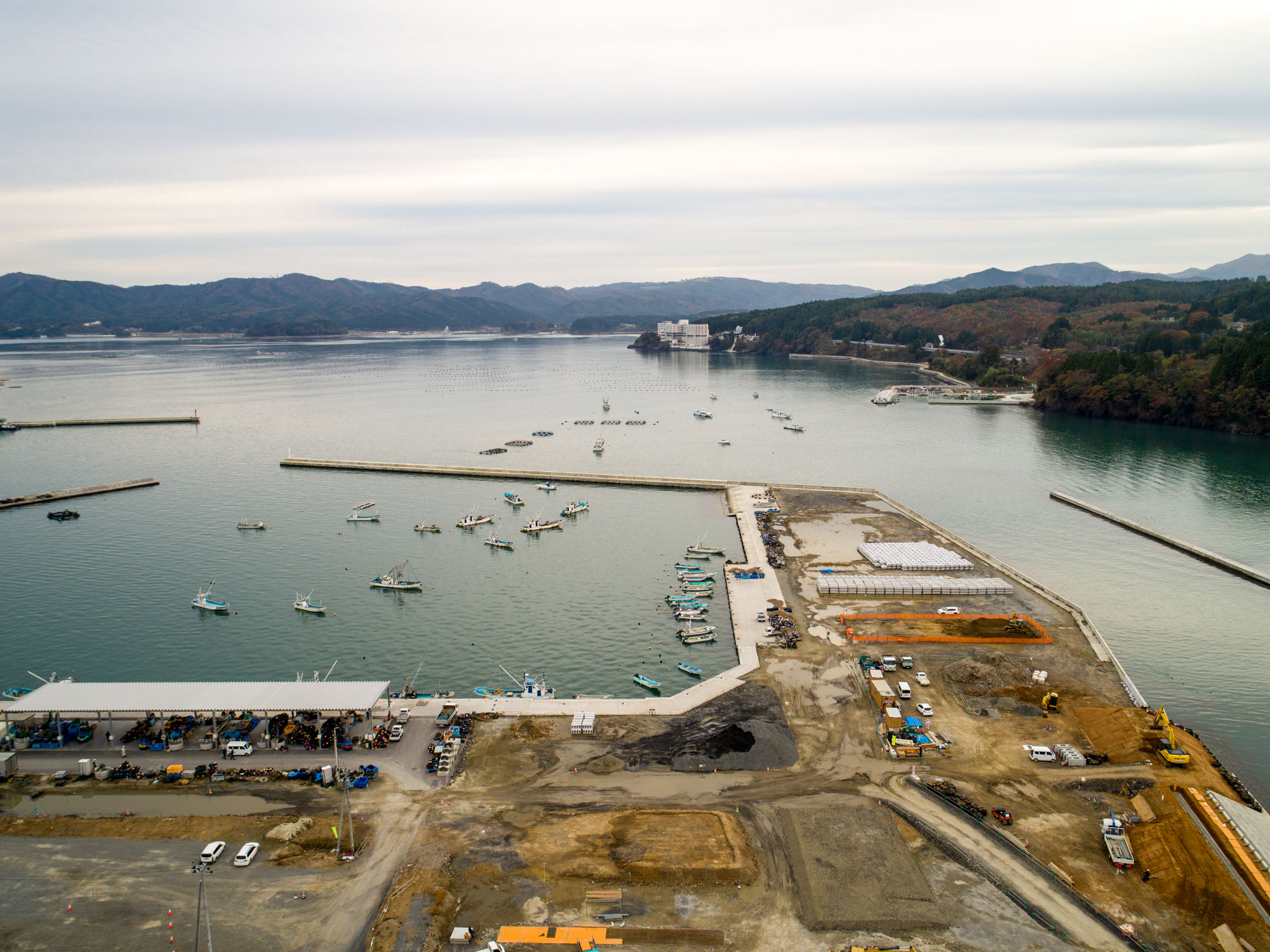 小型の船が複数浮かぶ海辺の港を上空から捉え、防波堤に囲まれた水辺の隣で建設作業が進み、背景には山岳地帯が広がる曇り空の下の写真