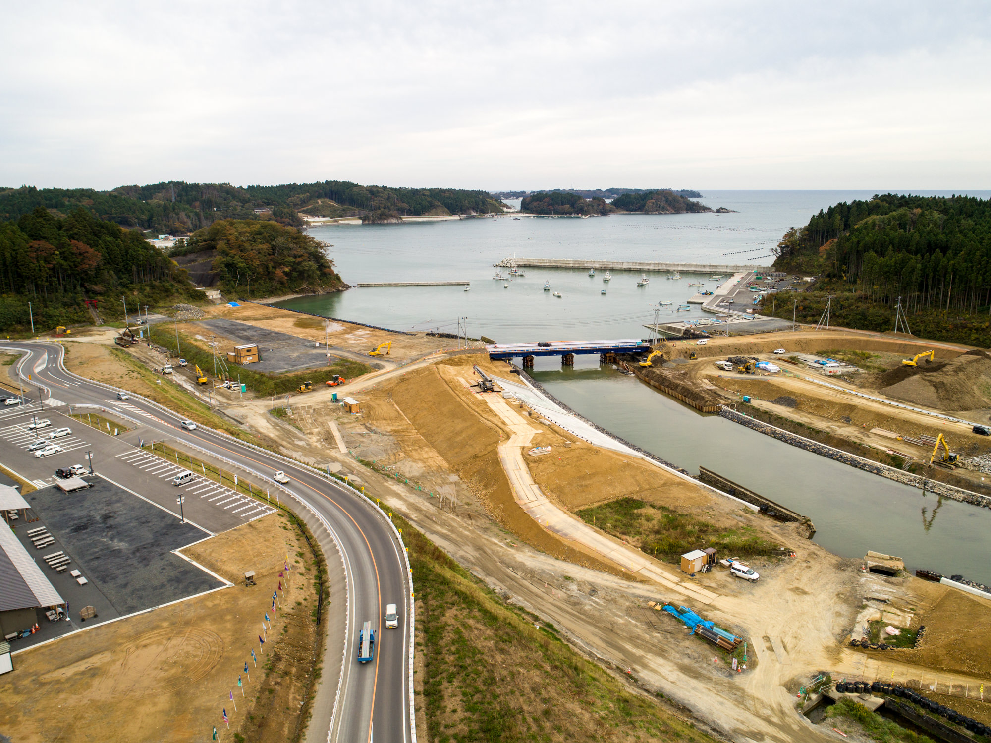 水路に架かる橋を中心とした海沿いの建設現場を捉え、周囲には道路や駐車場、森林が広がり、重機や車両が点在し、背景には海と小さな島々が曇り空の下に見える写真