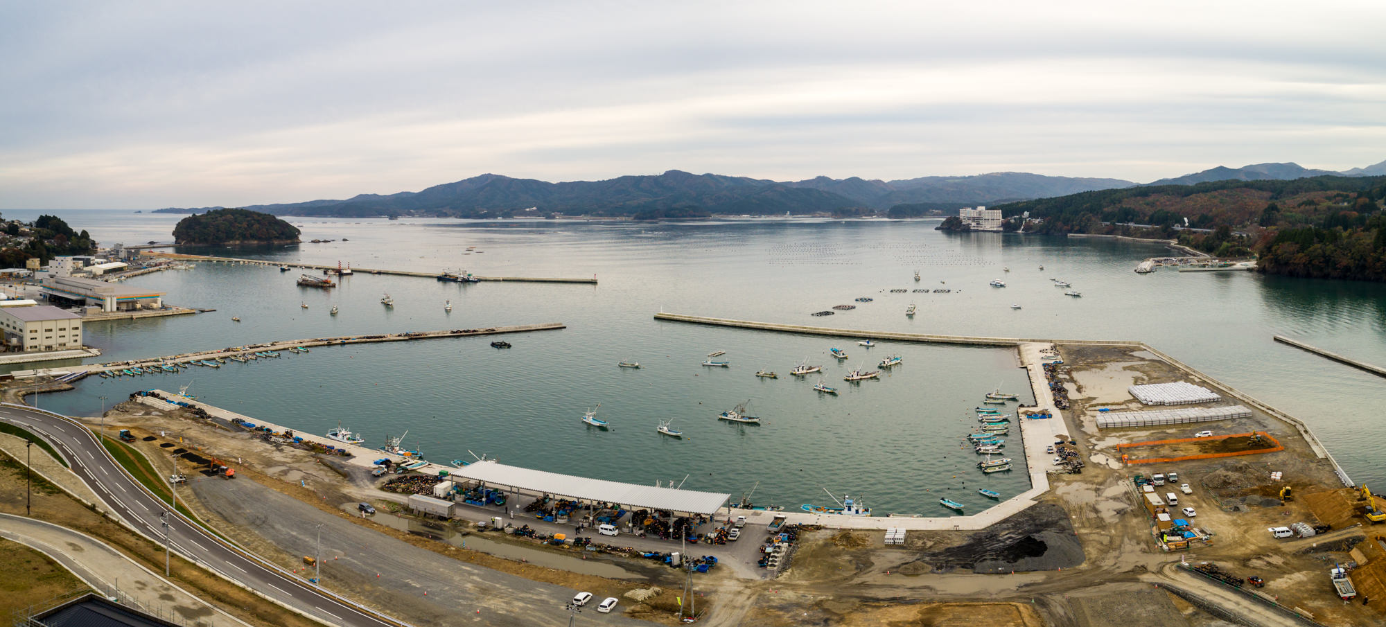 小型の船が多数停泊する港を中心に、防波堤や沿岸の建物、建設現場が広がり、背景には山々と遠くまで続く水面が見える曇り空の下の海辺のパノラマ写真