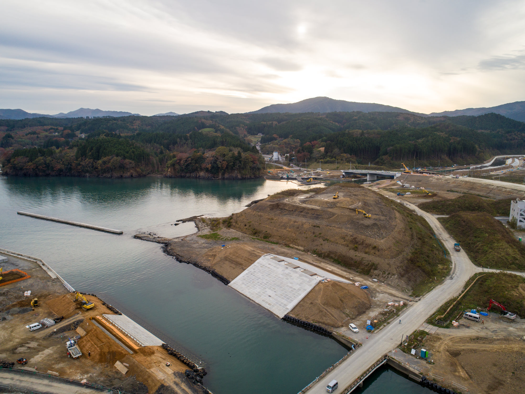 広大な水辺に面した海沿いの建設現場を捉え、道路や重機、コンクリート構造物が並び、背景には建設中の橋と森林に囲まれた丘が広がる、雲間から光が差す曇り空の下の写