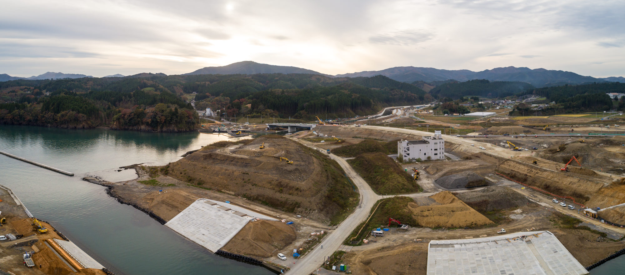 水辺の近くに広がる建設現場をパノラマで捉え、大型のコンクリート構造物や道路、重機が点在し、背景には丘と山々が広がる、緑がまばらな風景の写真