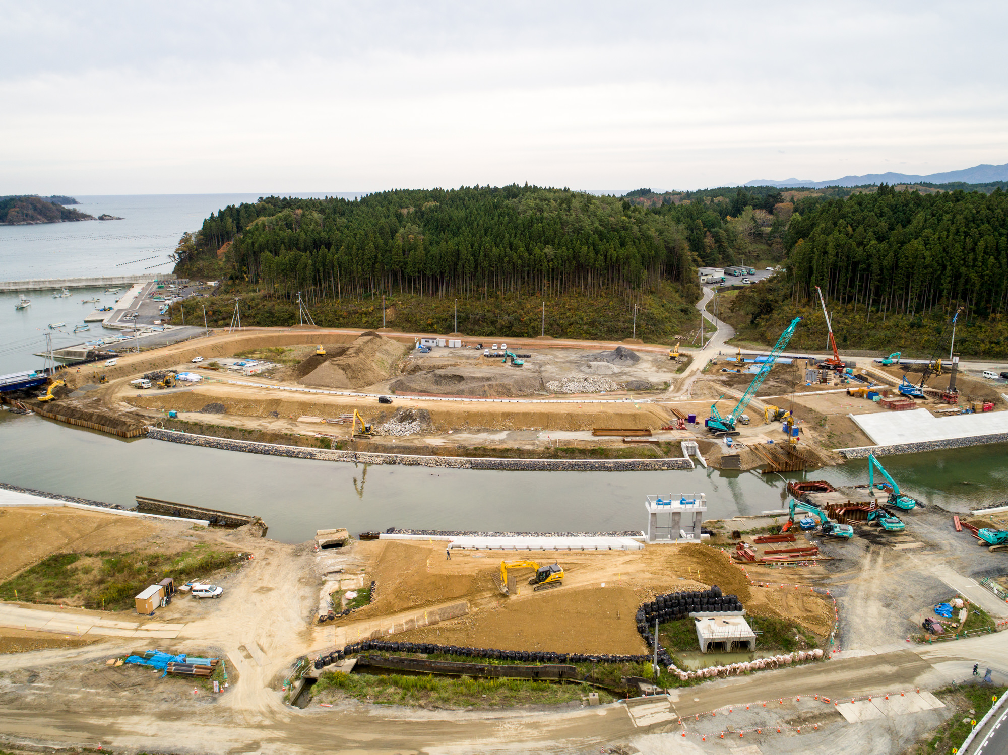 海へと続く川沿いの広大な敷地で、重機や設備が点在する建設現場を上空から捉え、周囲には森林が広がり、遠くには小島が浮かぶ海が見渡せる曇り空の下の写真