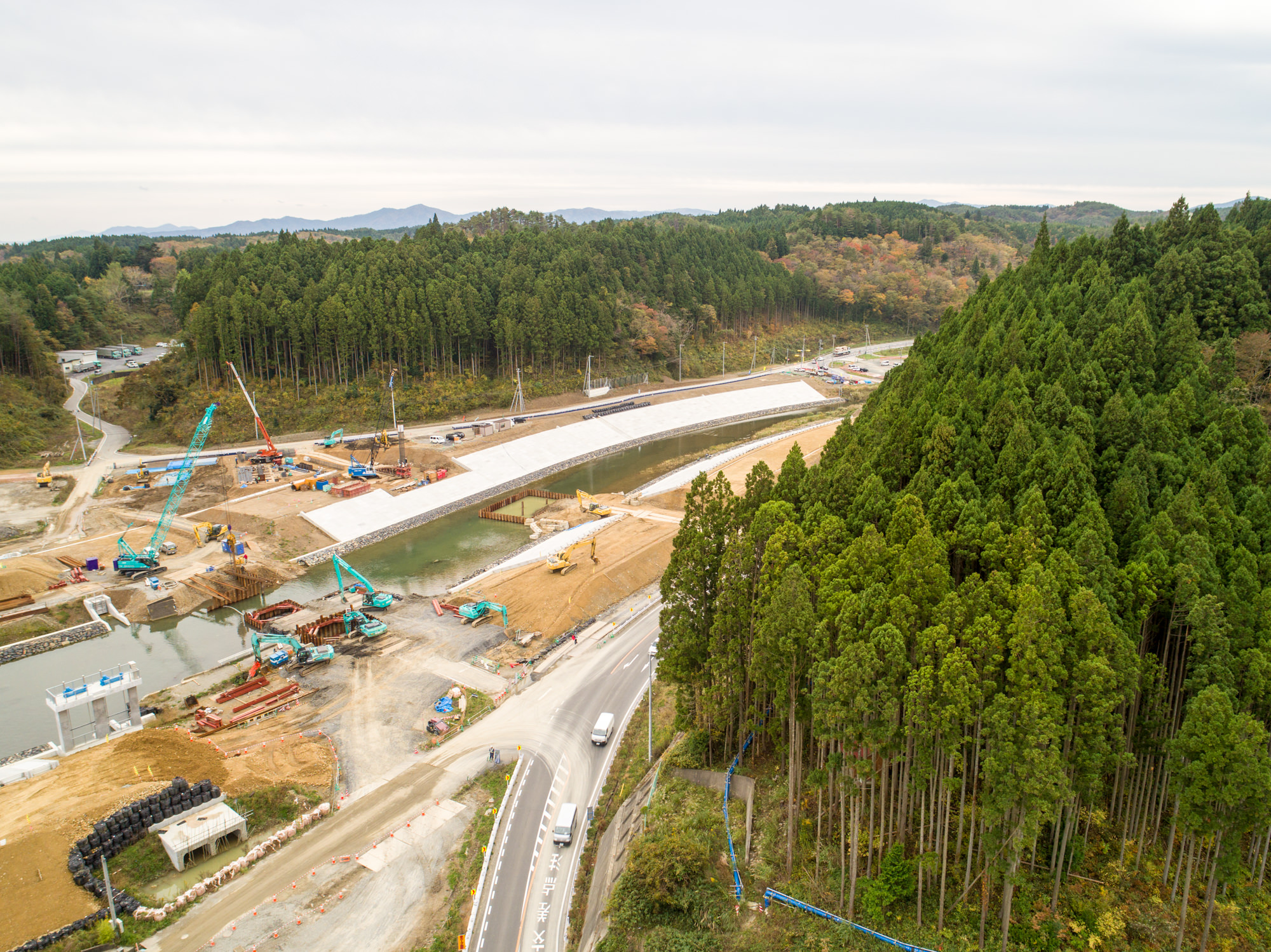 森林に囲まれた地域でクレーンや重機が川に架かる橋の建設を進めており、周囲には密集した木々や曲がりくねった道路、遠くに丘が広がる曇り空の下の写真