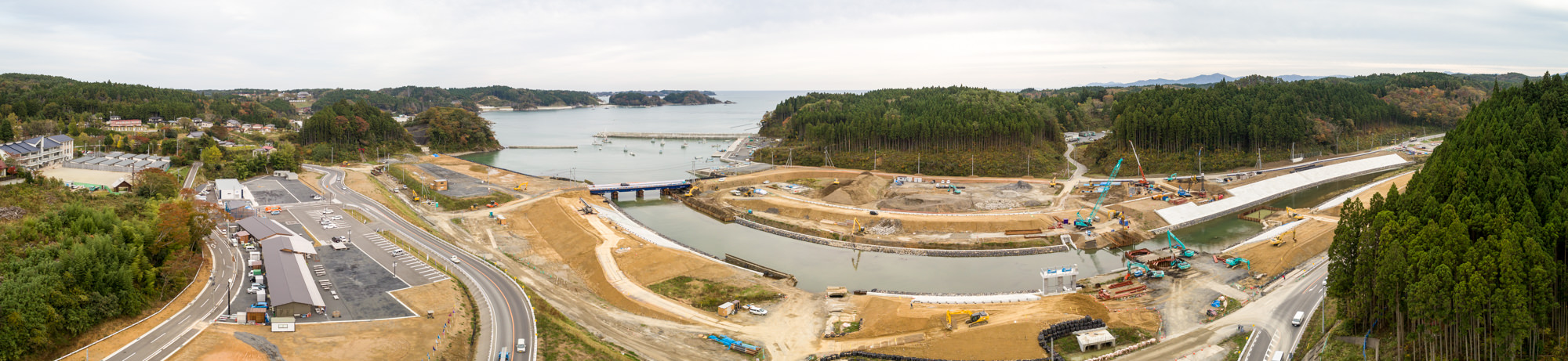 川が流れる海沿いの建設現場をパノラマで捉え、クレーンや重機、道路、建物が点在し、周囲には森林と丘が広がり、背景には海が見える写真