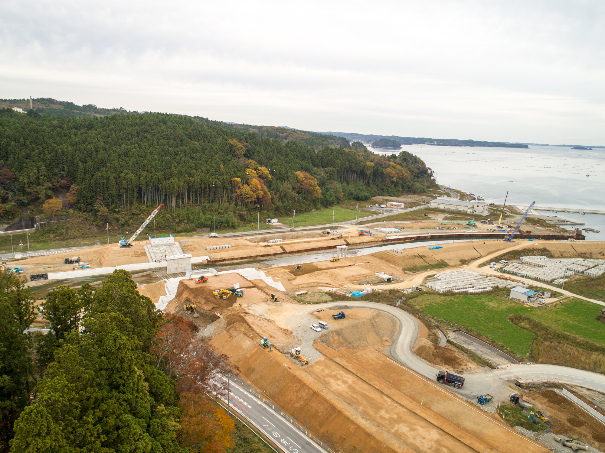 森林に囲まれた丘の近く、海辺に位置する広範囲な建設現場を上空から捉え、さまざまな重機や車両が点在する様子が見える写真