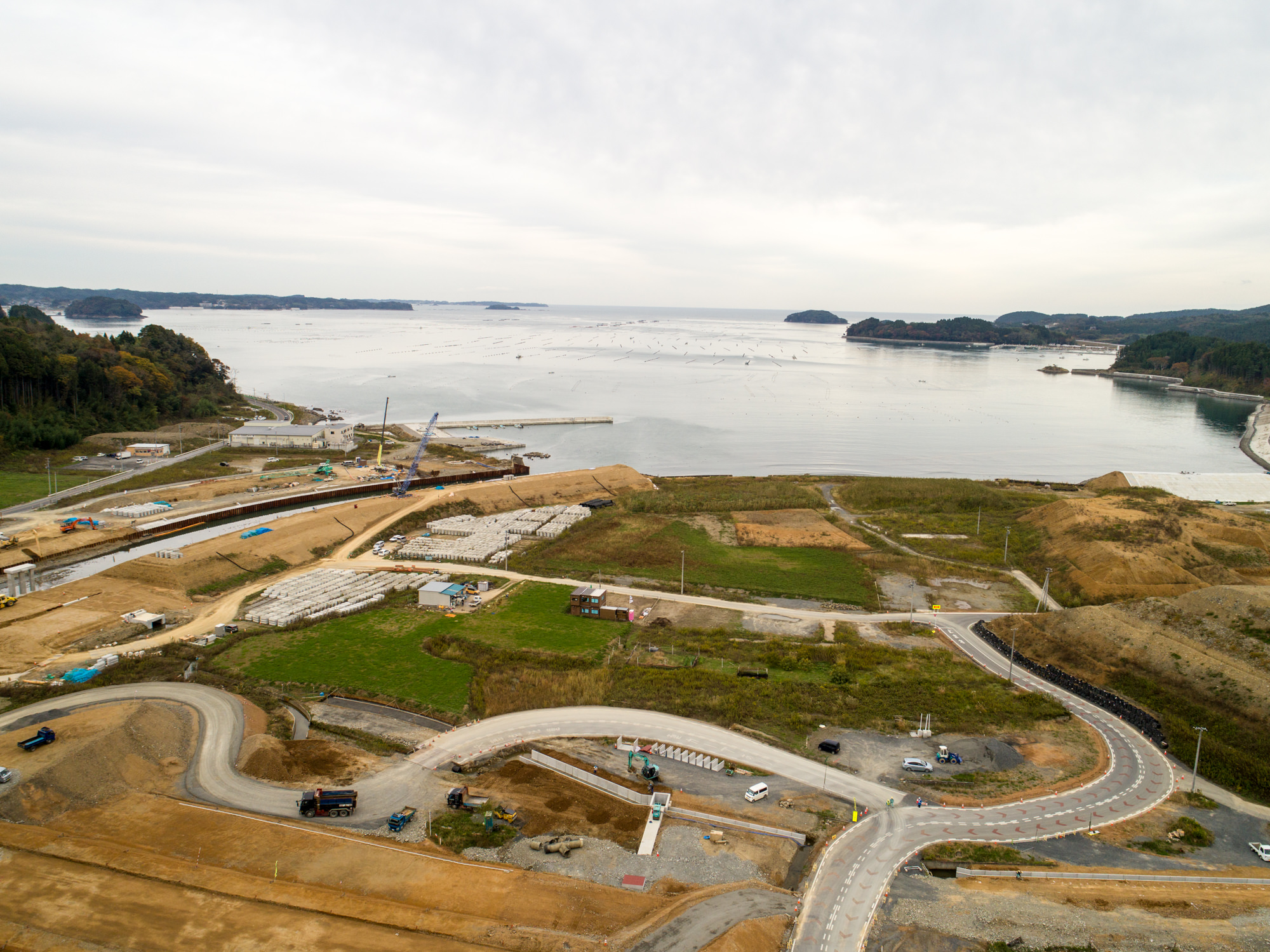 海を背景にした建設現場で、資材や車両、重機が点在し、草地と土の道が入り混じり、小さな島々が穏やかな海に浮かぶ曇り空の下の写真