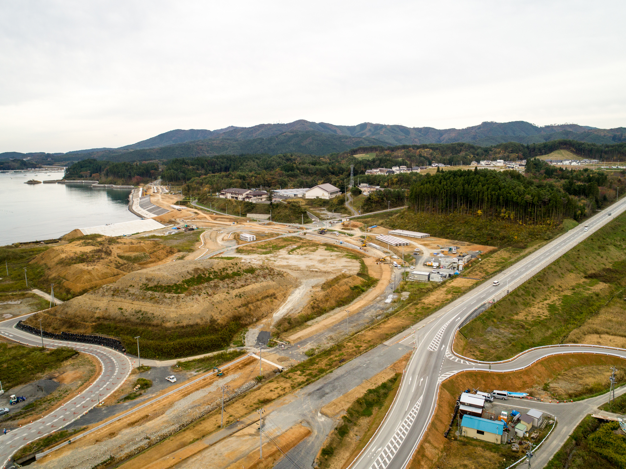 海岸沿いの広大な土地に曲がりくねった道路や建設現場が広がり、遠くには建物と木々に覆われた丘が見える曇り空の下の写真