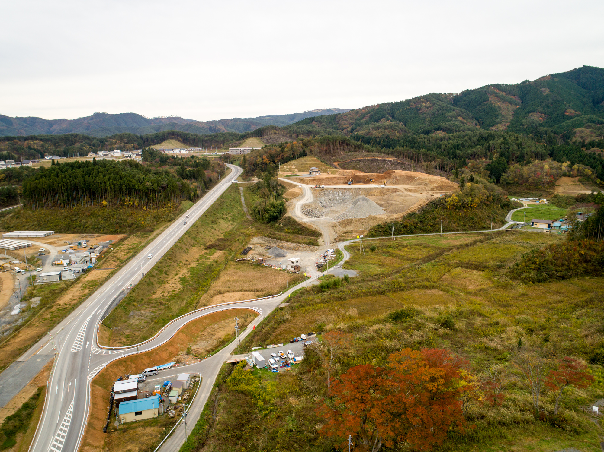 緑豊かな戸倉地区の建設現場を空から撮影した写真で、山々を背景に曲がりくねった道路が建設現場へと続き、整地された土地や重機、小さな建物と車両が点在する風景の写真