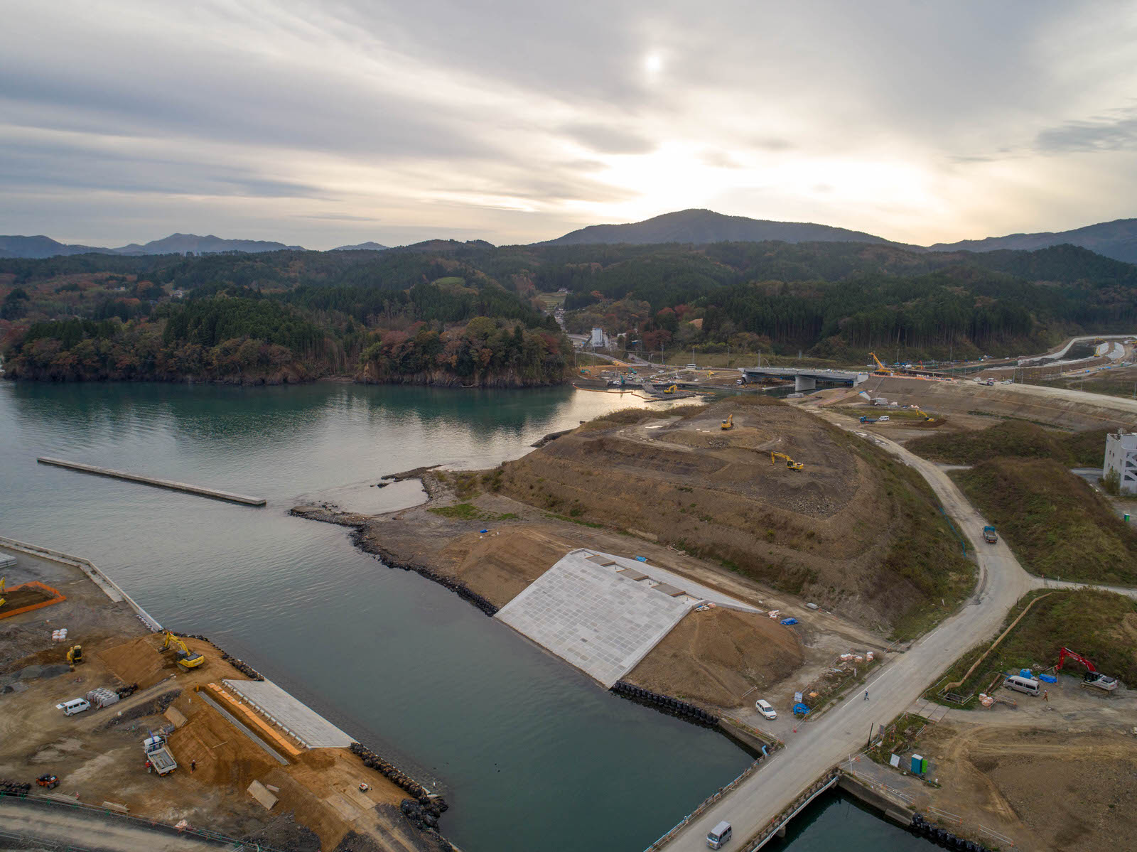 水辺近くの建設現場を空から撮影したもので、複数の道路や通路が敷地内を走り、車両や建設機材が点在し、広い土地が造成中で、背景には木々に覆われた丘や山が広がり、曇り空の中に日差しが差し込む写真