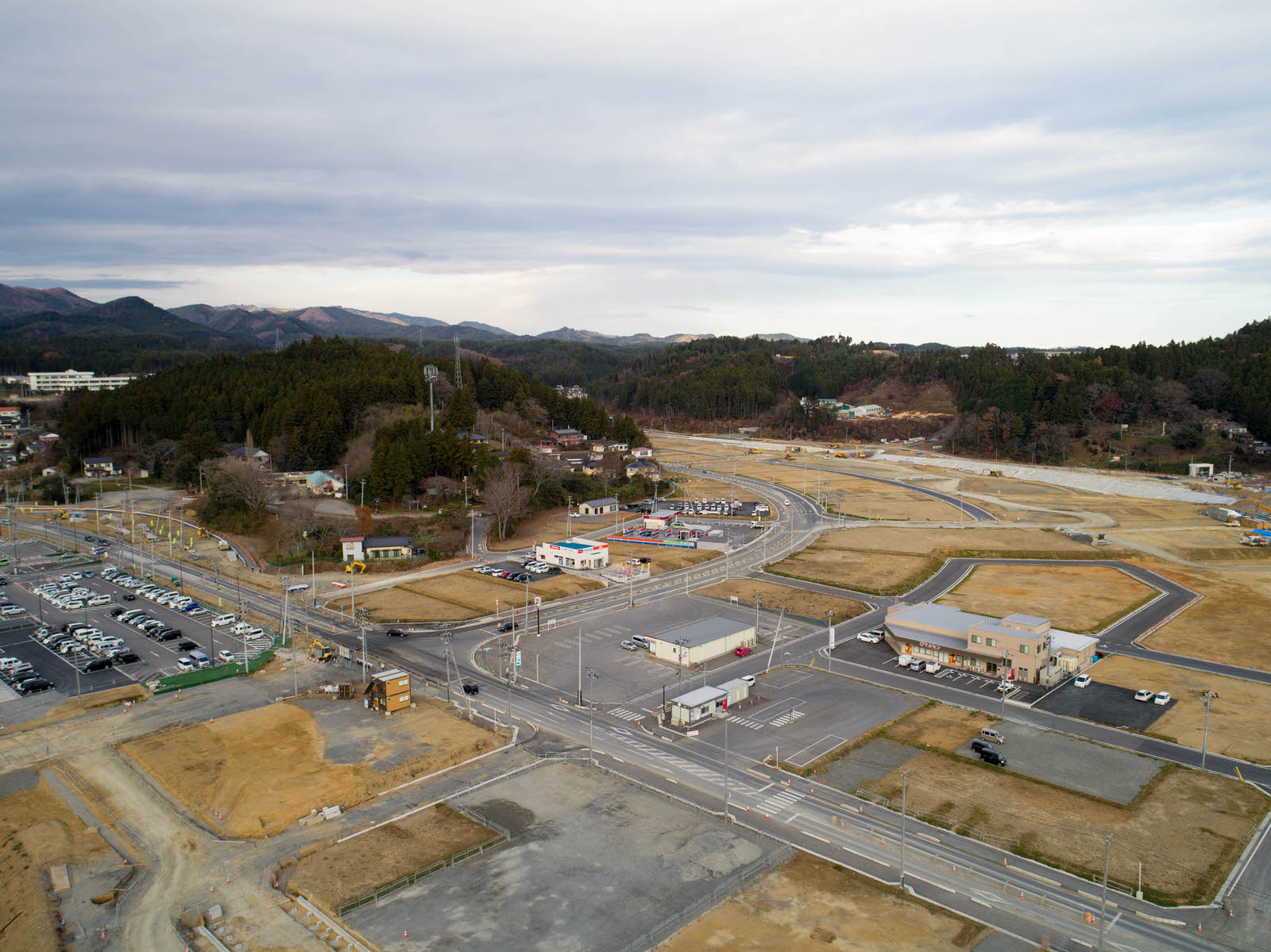 開発途中の土地と建物が混在する志津川地区を空から撮影したもので、複数の建物や道路、駐車場が見られ、草地や土の部分が広がり、一部は工事中の様子を示し、背景には木々に覆われた丘があり、曇り空が広がる写真