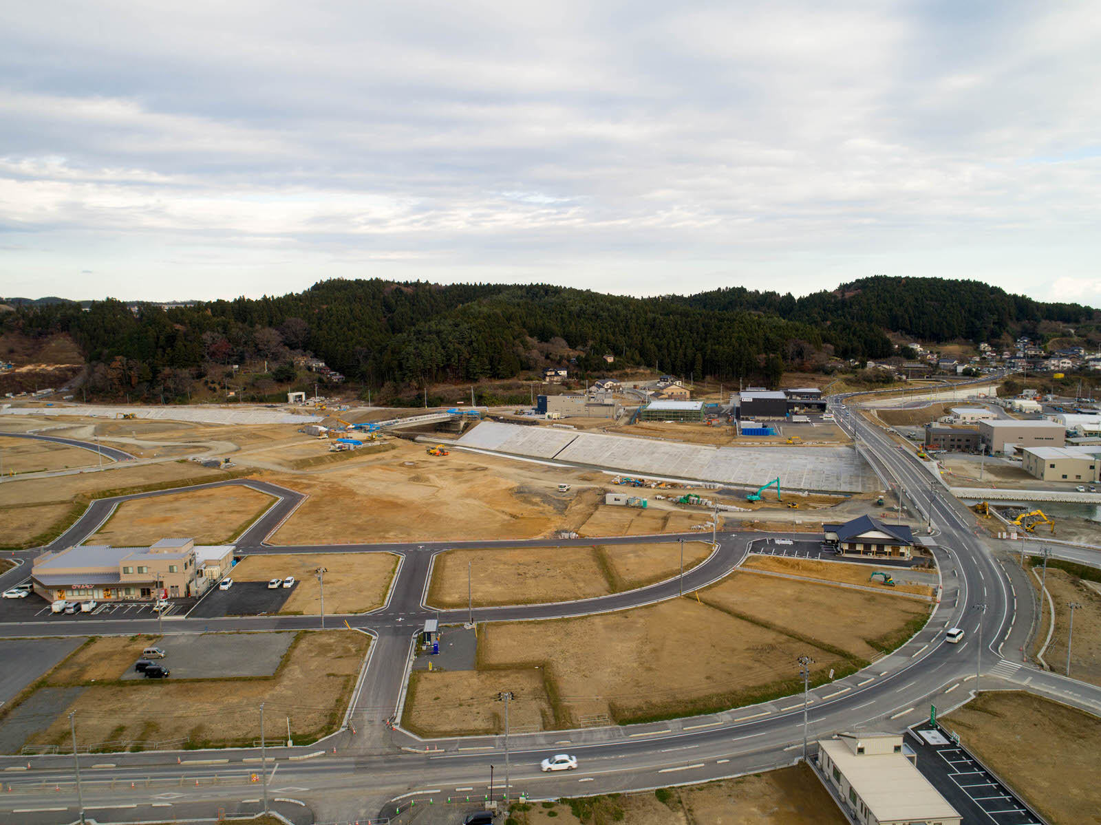 複数の道路と建物が点在する建設現場を空から撮影したもので、開発初期段階の様子がうかがえ、すでに完成した構造物と建設中のものが混在し、重機や建設機材が見られ、背景には森林に覆われた丘と曇りがちの空が広がる写真