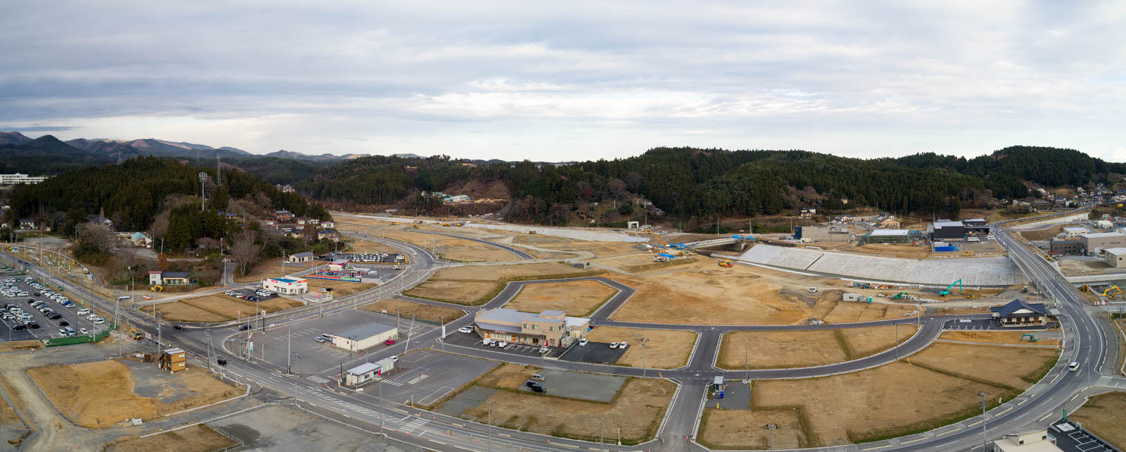 広い造成地を高台から撮影したもので、複数の道路と建物が点在し、地面の一部はむき出しで建設機材が見られ、背景には木々に覆われた丘陵地が広がり、曇り空が広がるパノラマ写真