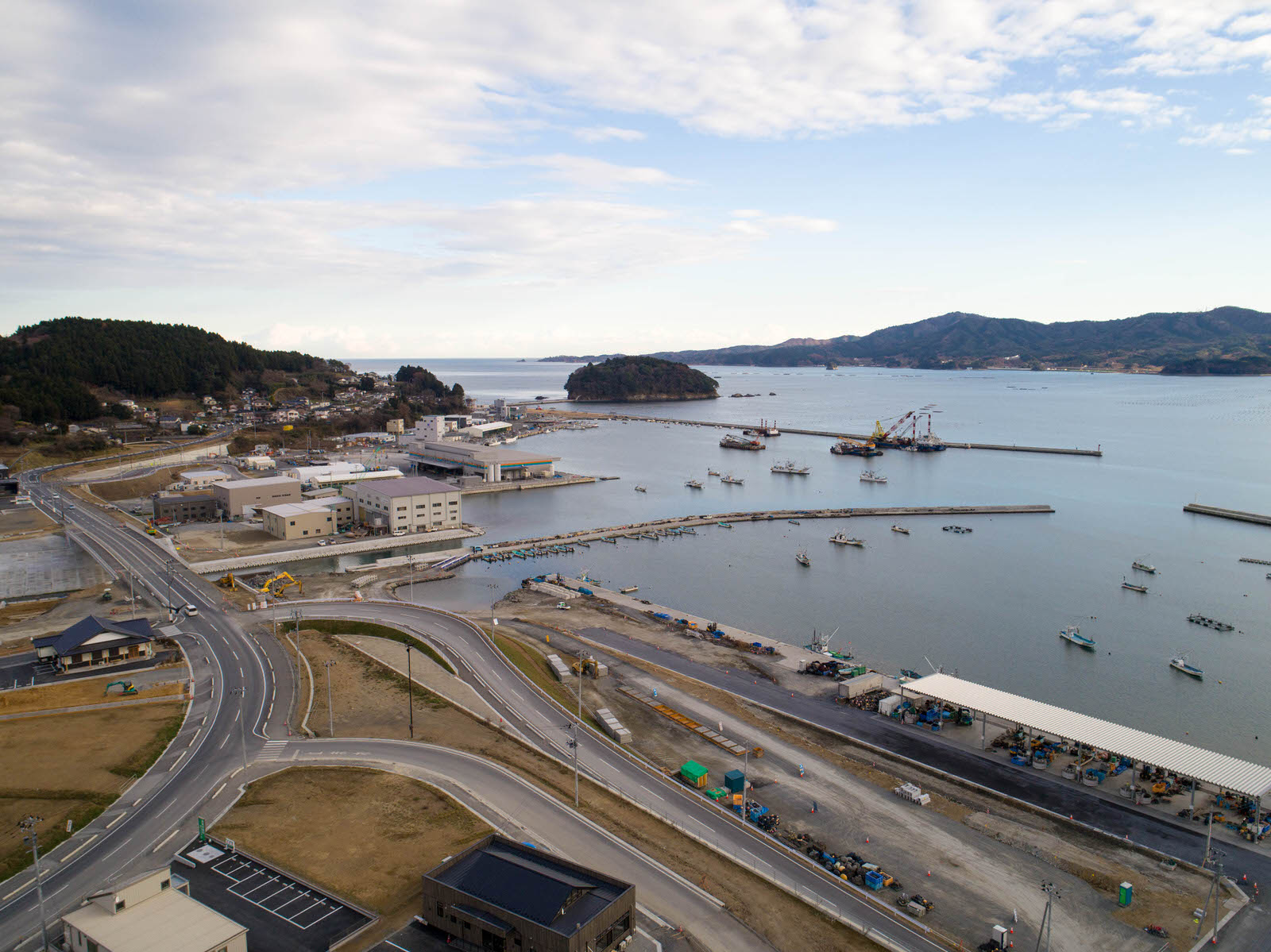 港のある海沿いの地域を空から撮影したもので、岸辺には倉庫などの工業施設と思われる建物が並び、港内には複数の防波堤と船が停泊し、周囲には道路や緑地、背景には丘陵地が広がり、青空が部分的に見える曇り空の写真