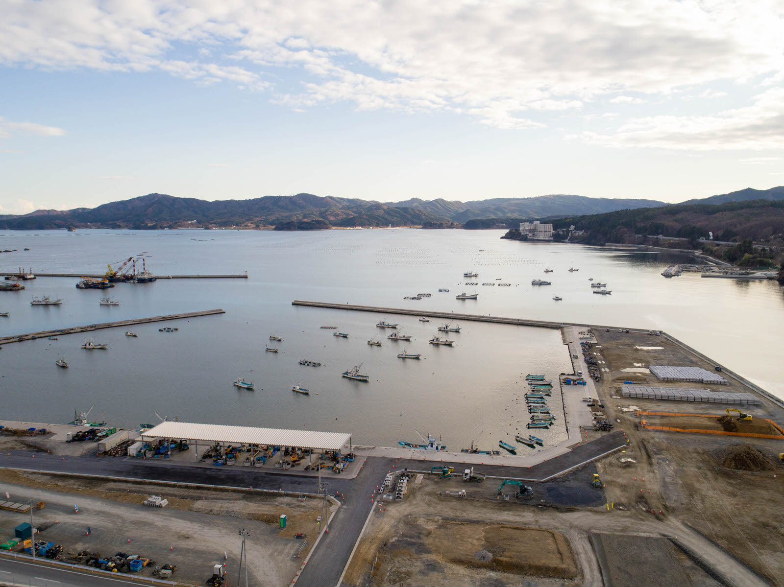 港のある海沿いの地域を空から撮影したもので、水面には複数の船が停泊し、岸辺には大きな屋根付きの施設を含む構造物が並び、背景には山々と部分的に雲がかかった空が広がる写真