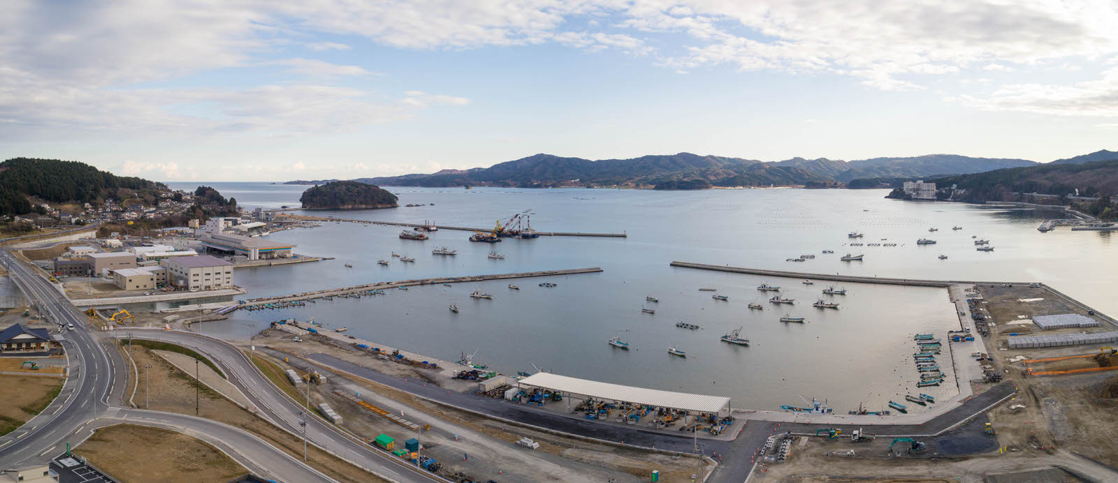 港のある海沿いの志津川地区を空から撮影したもので、水面には複数の船が停泊し、岸辺には建物や構造物が並び、背景には丘や山が広がり、曇りがちの空の下で海岸付近では開発工事が行われているパノラマ写真