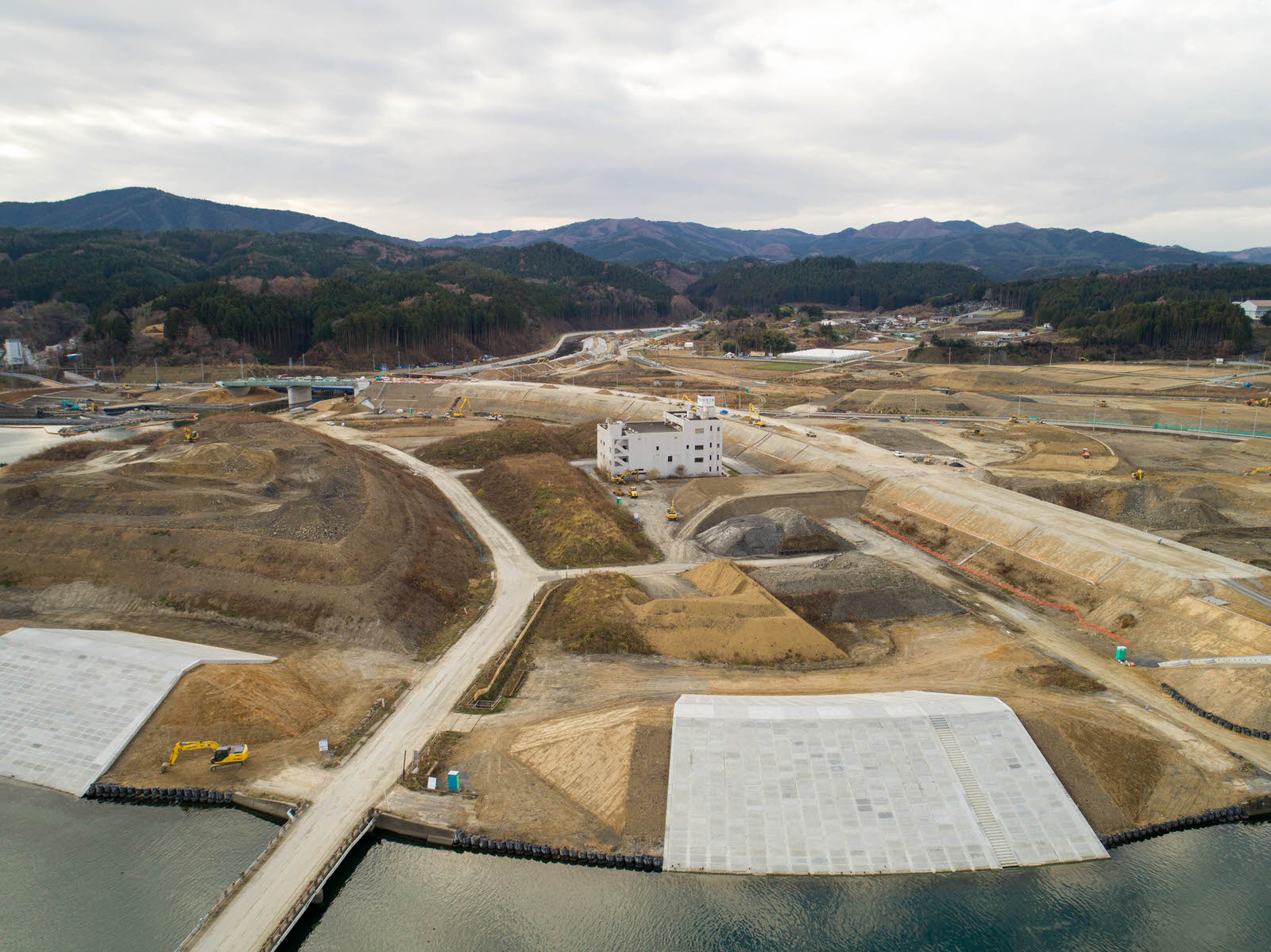 土木工事が進む建設現場を空から撮影したもので、中央には白い外観の建物があり、周囲には土の盛り上がりや重機、複数の未舗装道路が広がり、手前には水辺とコンクリート構造物が見られ、背景には森林に覆われた丘や山が曇り空の下に広がる写真