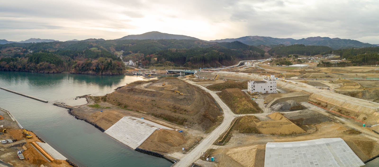 水辺に近い建設現場を空から撮影したもので、敷地内には複数の道路や通路が走り、土の盛り上がりやコンクリートで覆われた部分が点在し、背景には山や森林が広がり、曇り空が広がるパノラマ写真