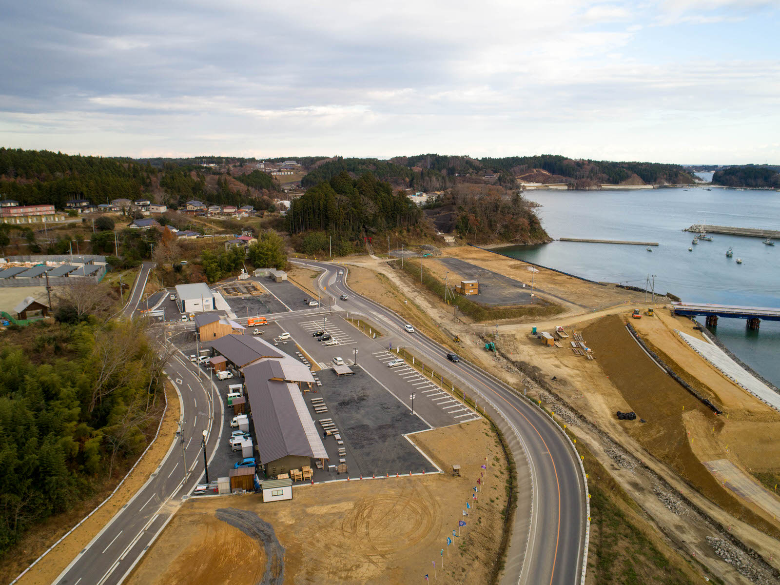 海に面した平地は造成中で、道路を挟み広い駐車場がある商業施設の建物があり周りに森が点在している風景を上空から広く撮影した写真
