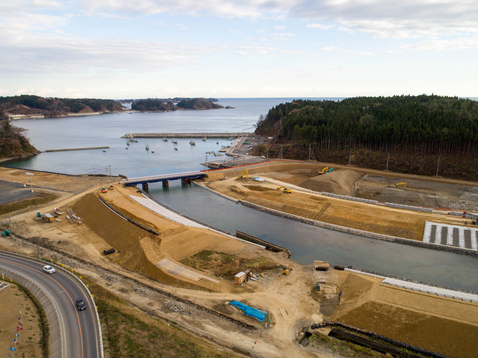 川沿いの建設現場を空から撮影したもので、水路には橋が架かり、両岸には重機や建設機材が点在し、周囲には森林と造成中の土地が広がり、水路の出口付近には船が停泊している写真