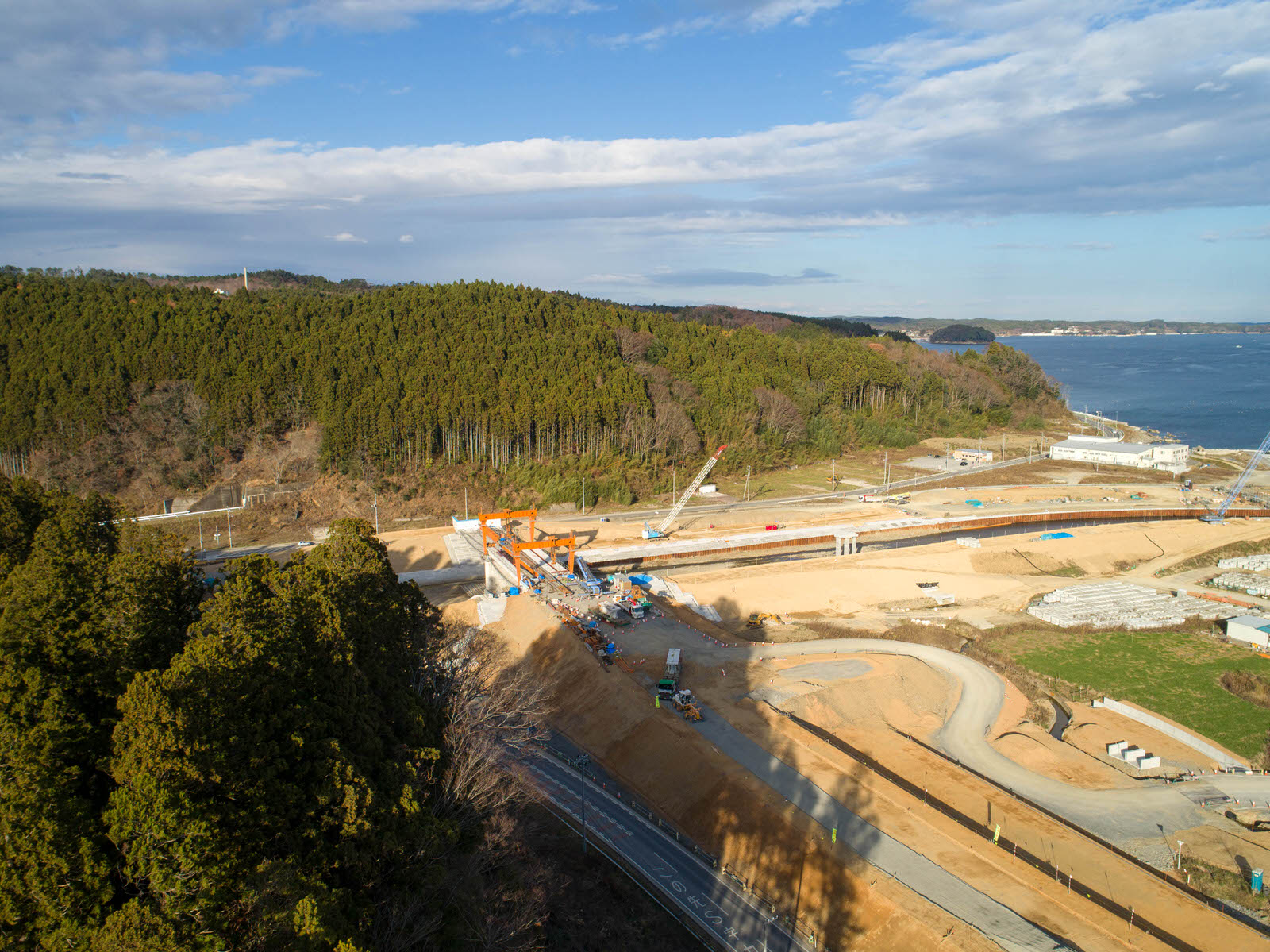森林に囲まれた建設現場を空から撮影したもので、大型クレーンや重機が高架の建設作業を行っており、背景には水辺が広がり、青空が部分的に見える曇り空の写真