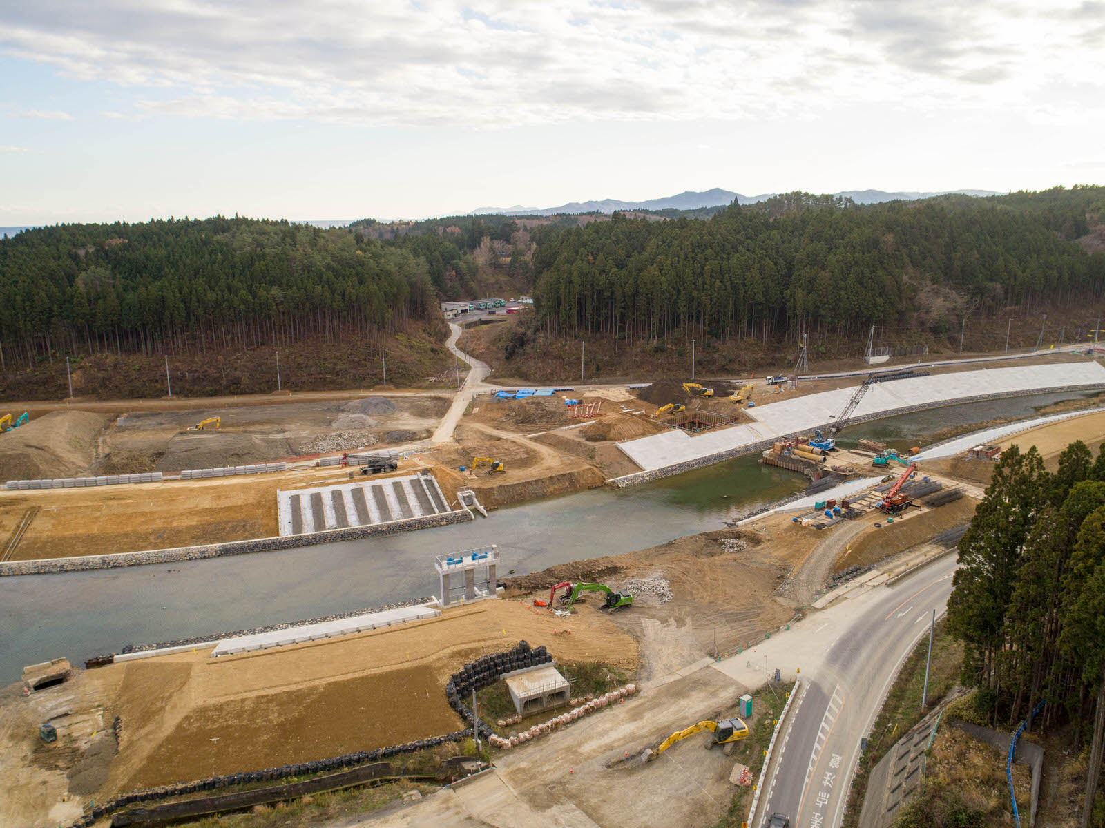 川沿いの建設現場を空から撮影したもので、周囲は森林に囲まれ、敷地内にはショベルカーやクレーンなどの重機が点在し、川岸の補強工事と思われるコンクリート構造物が見られ、道路が現場沿いに走り、奥の森林地帯へと続いている写真