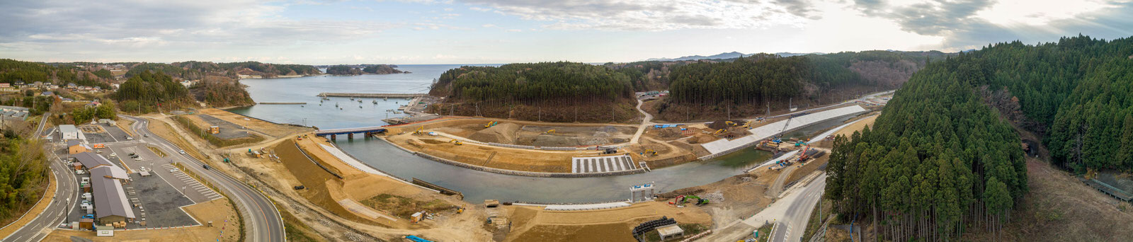 川が中央を流れる海沿いの地域を広く空から撮影したもので、川には橋や道路が架かり、左側には建物や駐車場、右側には密集した森林が広がり、背景には海と森林に覆われた丘陵地が見え、川沿いや周辺でインフラ整備が進められているパノラマ写真