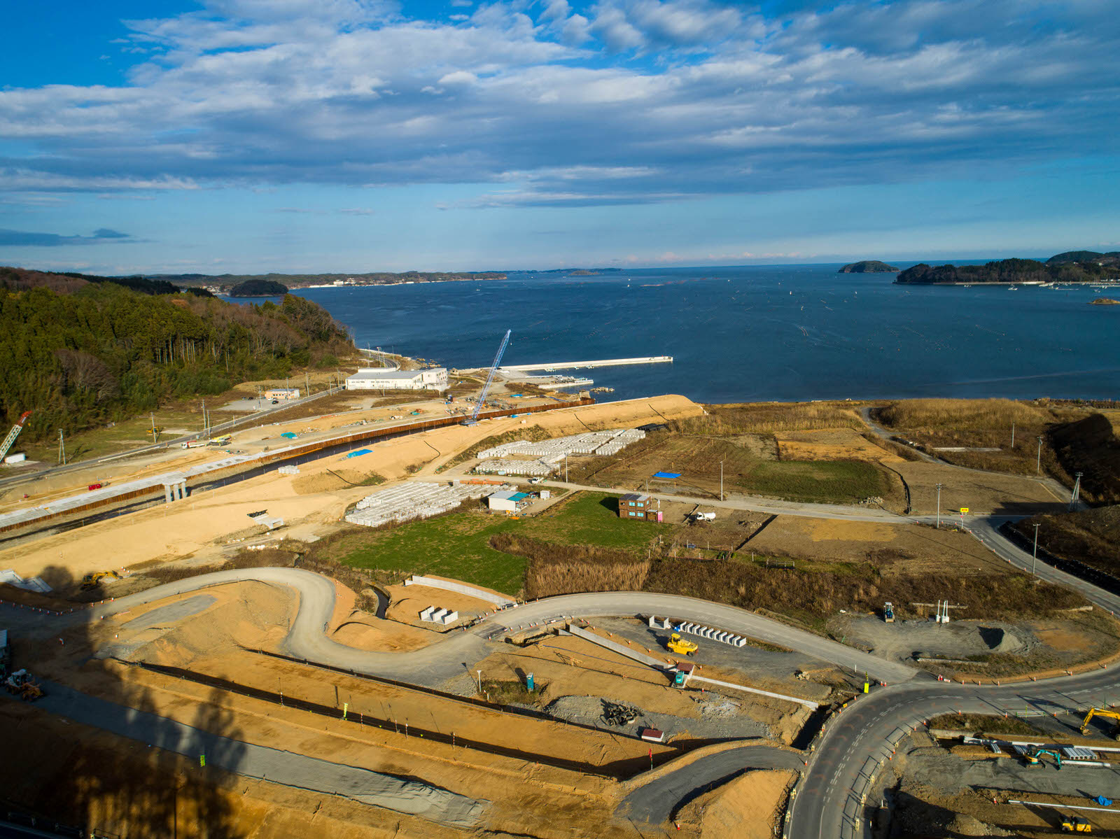 海沿いの建設現場を空から撮影したもので、敷地内には建設資材や重機、建設途中の構造物が点在し、曲がりくねった道路や舗装・未舗装の地面が広がり、背景には桟橋が突き出た水辺と木々や丘陵地が見える写真