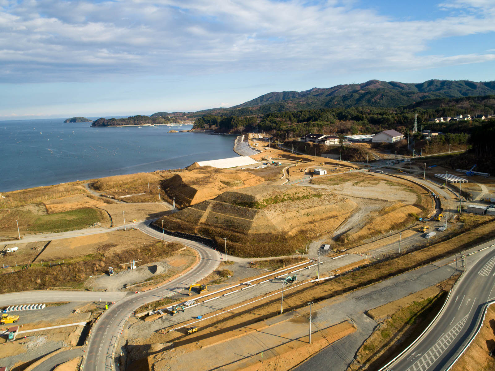海沿いの建設現場を空から撮影したもので、湾曲した海岸線に沿って道路が走り、重機や土砂の山が点在する広範な造成地が広がり、背景には船が浮かぶ海と、内陸には木々に覆われた丘や山が見える写真