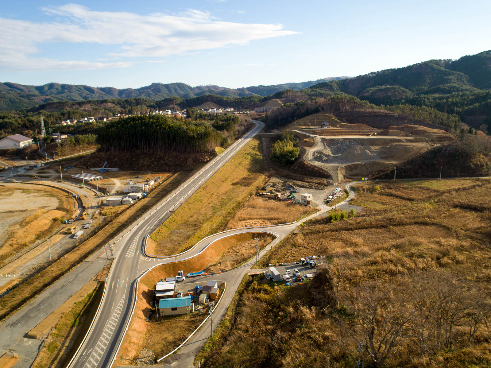草地と建設現場に囲まれた戸倉地区の風景を空から撮影したもので、中央には複数車線の新しい道路が通り、背景には森林を含む丘や山が広がり、道路沿いには建物や車両が見られる晴天の写真