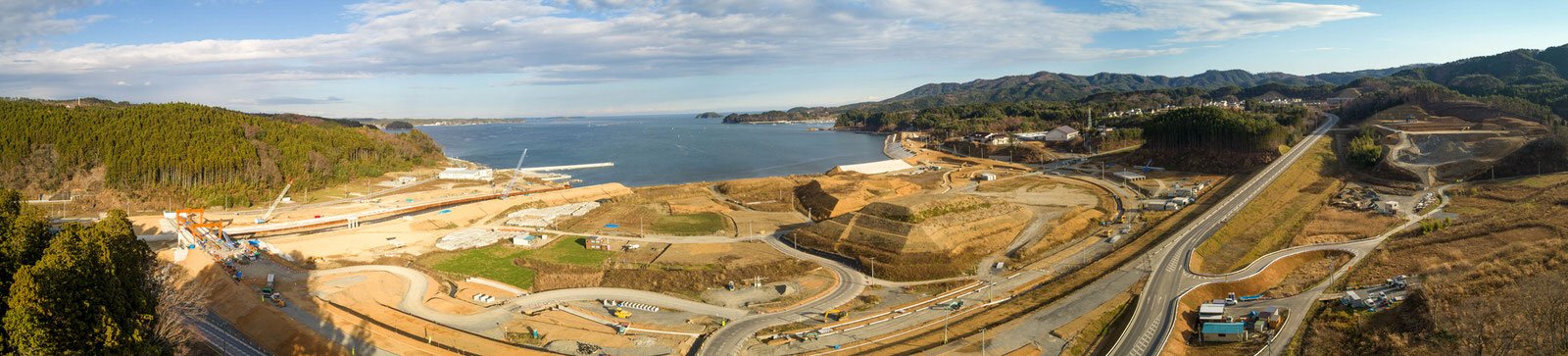 海沿いの建設現場を広く見渡した空撮写真で、湾曲した道路や掘削された土地、建設中の構造物が点在し、左奥には森林、右奥には山岳地帯が広がり、青空が部分的に見える曇り空のパノラマ写真
