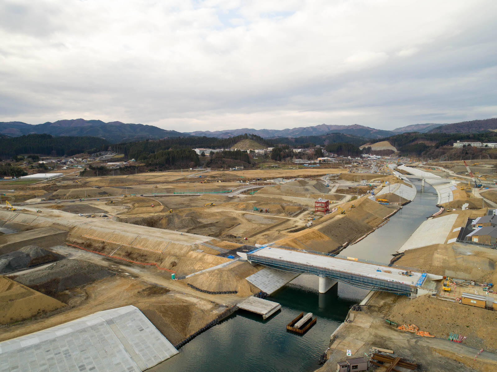 曇り空が広がるなか、川に架かる建設途中の橋を中心にした現場を高台から撮影したもので、周囲には土木作業や道路、重機が広がり、背景には山々が連なる建設現場の写真