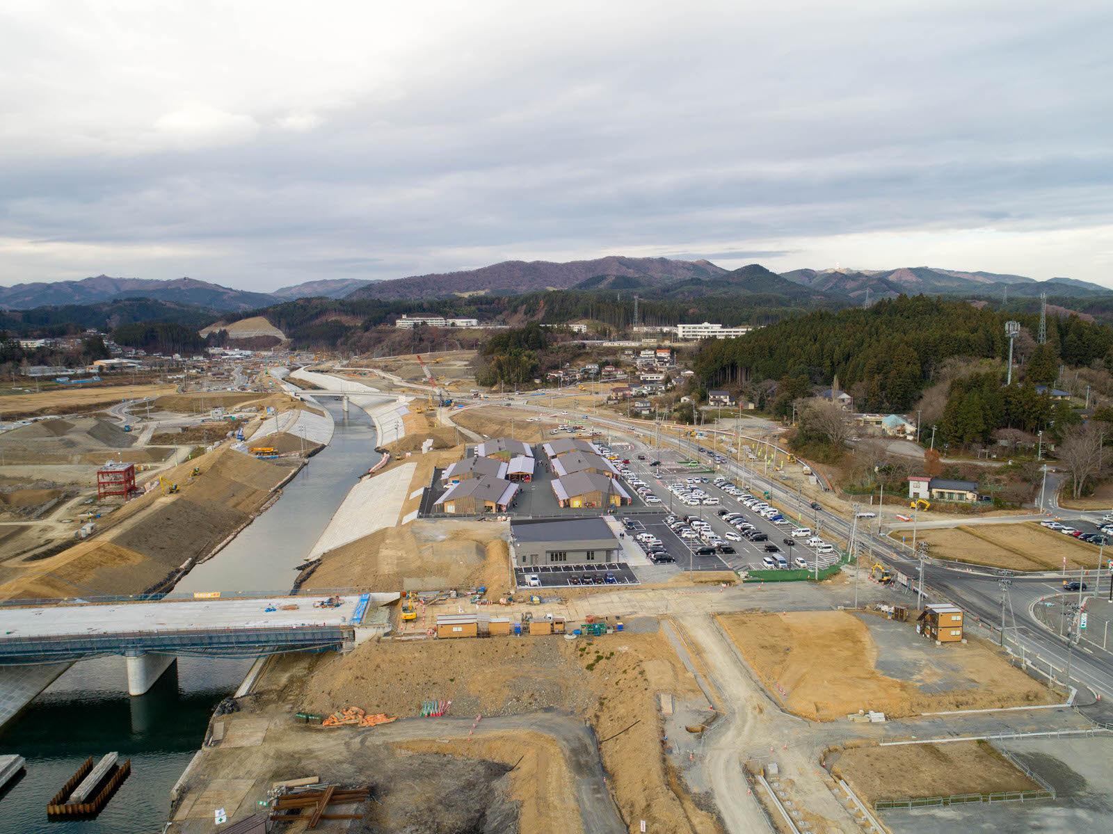川沿いの建設現場を空から撮影したもので、橋が建設中であり、背景には建物や構造物が点在し、すでに整備された道路や駐車場が見られ、遠くには丘陵地と森林が広がる写真