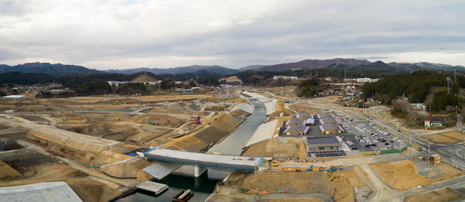 川が中央を流れる広大な建設現場を空から撮影したもので、複数の橋が建設中であり、川の両岸には建物や構造物が点在し、右側には車が並ぶ駐車場があり、背景には丘陵地と森林が広がる写真