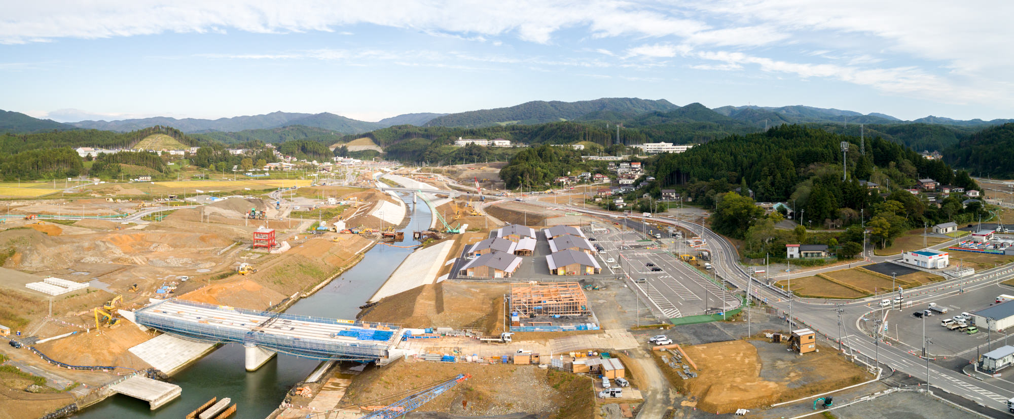川に架かる橋や道路、建物など複数の構造物が建設中で、周囲には丘や森林が広がり、資材や建設機器が点在する志津川地区の工事現場を広く見渡したパノラマ写真