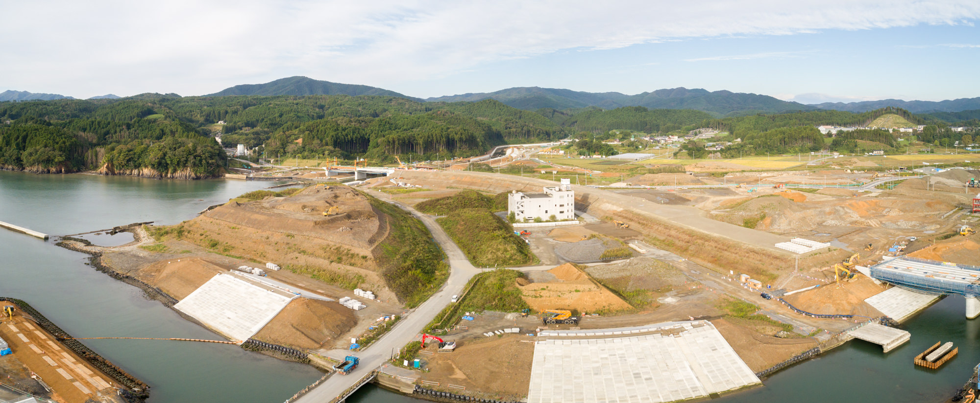 水辺近くの建設現場を広く見渡した風景で、広いコンクリート敷地や未舗装の道路、建設機器が点在し、背景には建物や構造物、さらに緑の丘や山々が広がる様子を、雲が浮かぶ空の下で上空から捉えた工事現場のパノラマ写真