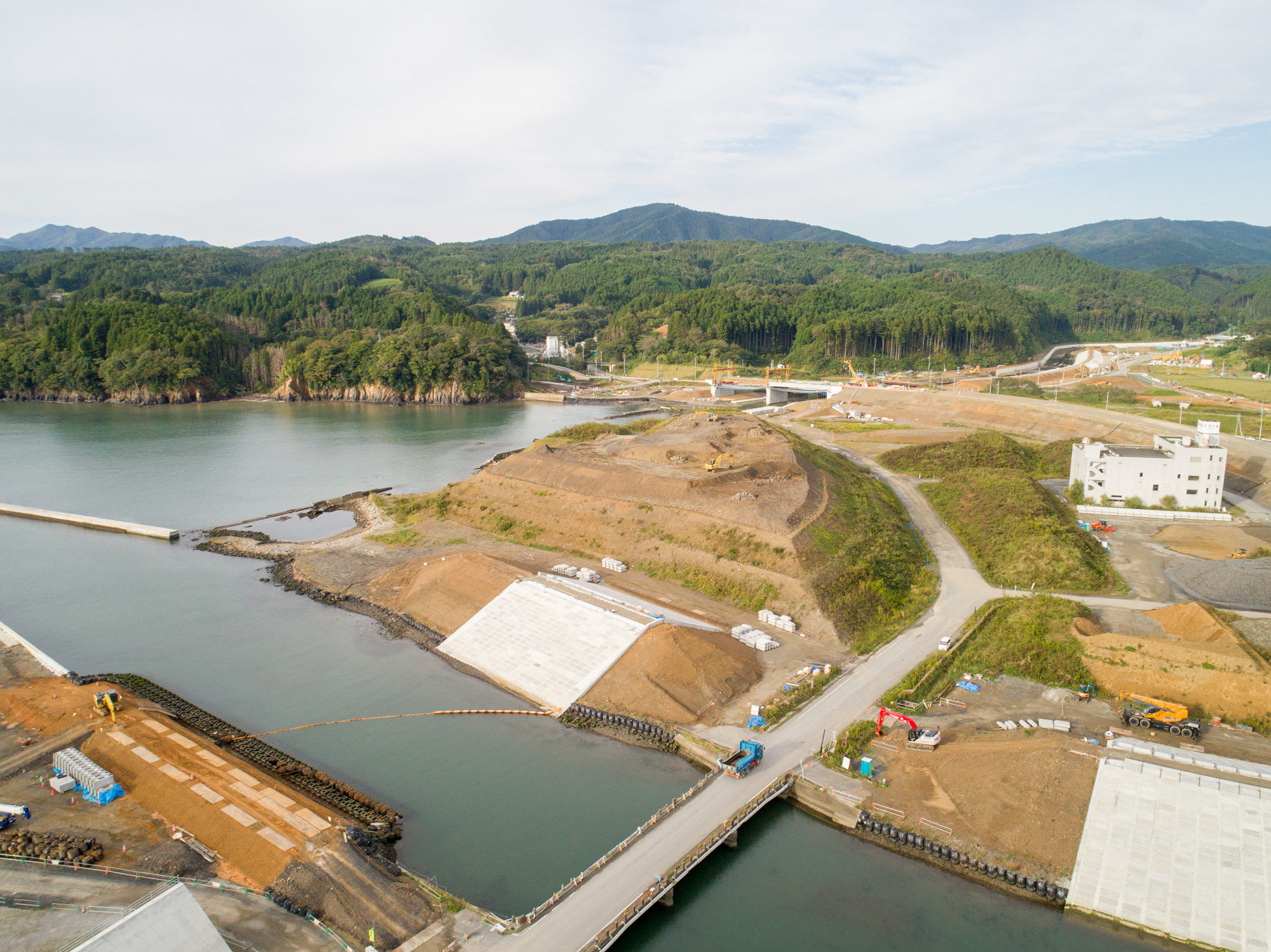川沿いの水辺の近くで、道路や橋、堤防が整備され、建設車両や機材が点在する工事現場を、周囲の木々に覆われた丘陵地とともに上空から捉えた写真