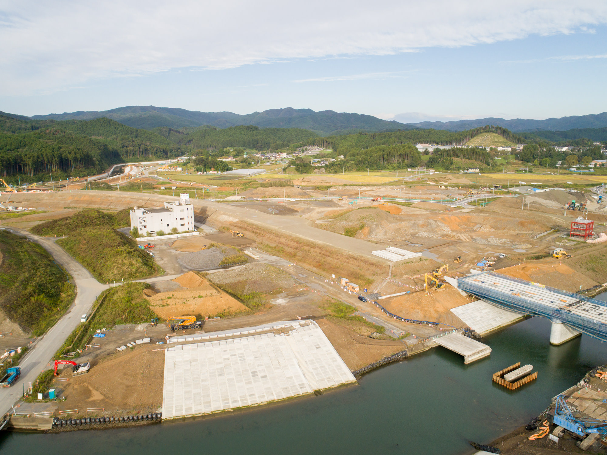 水辺に架かる途中の橋や大きなコンクリートの敷地、建設車両や機材、未完成の建物が点在し、周囲には丘陵地や森林、遠くに山々が広がる風景を上空から捉えた工事現場の写真