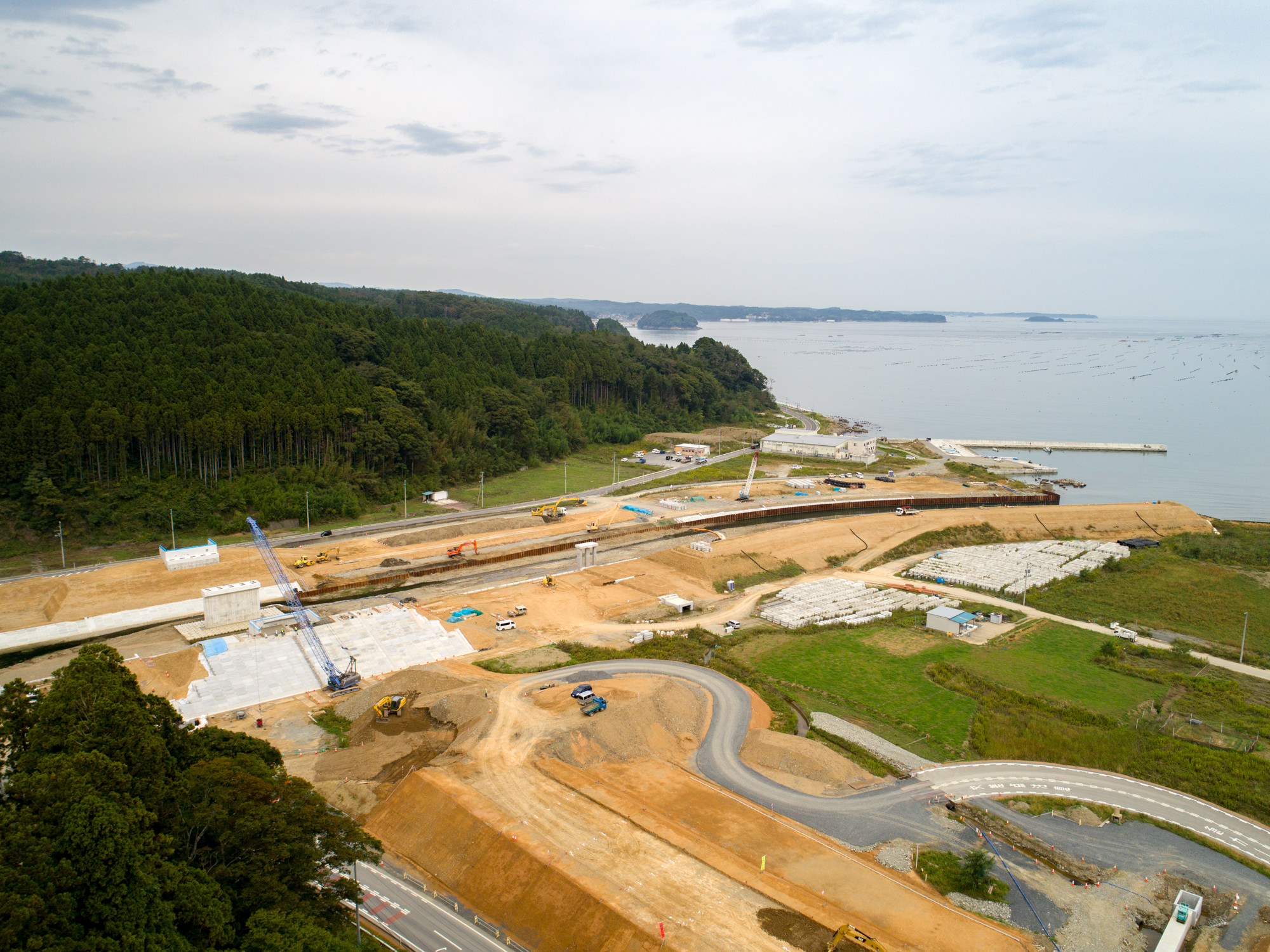 海に面した場所で、クレーンやショベルカーなどの建設車両が稼働し、複数の建物や道路が建設途中の様子を、片側に森林、もう片側に海を望む環境の中で捉えた工事現場の写真