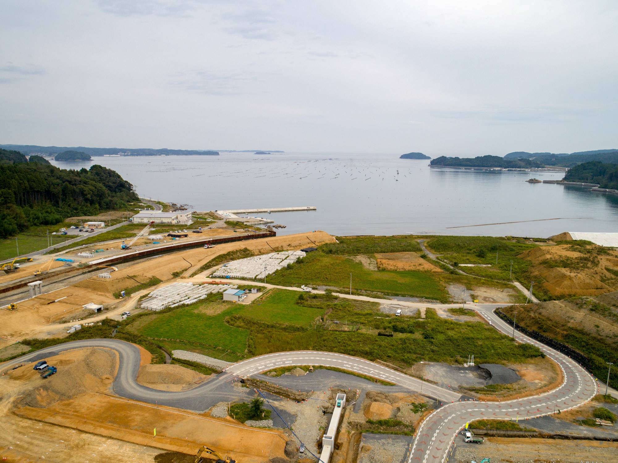 穏やかな湾と遠くに小さな島々を望む海岸沿いに、曲がりくねった道路や複数の建設現場、緑地が点在する開発中の地域を上空から捉えた沿岸風景の写真