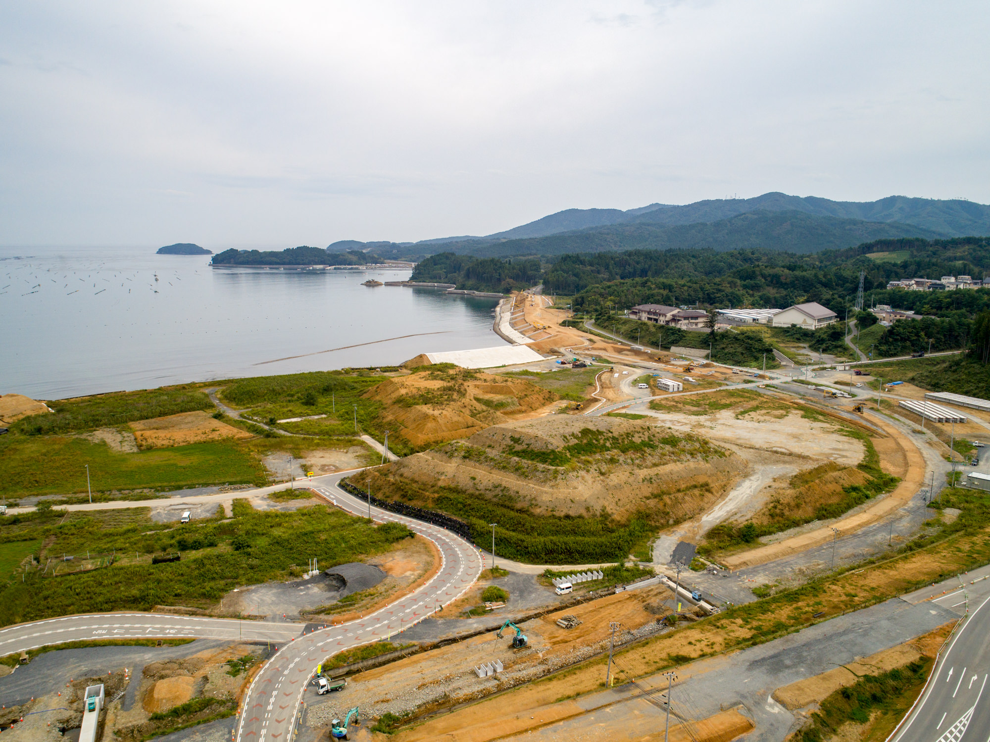 手前に曲がりくねった道路と建設機械が見え、緑地が点在する中、砂浜が海へと続き、背景には丘や山々が広がる自然と開発が混在した沿岸地域を上空から捉えた風景の写真