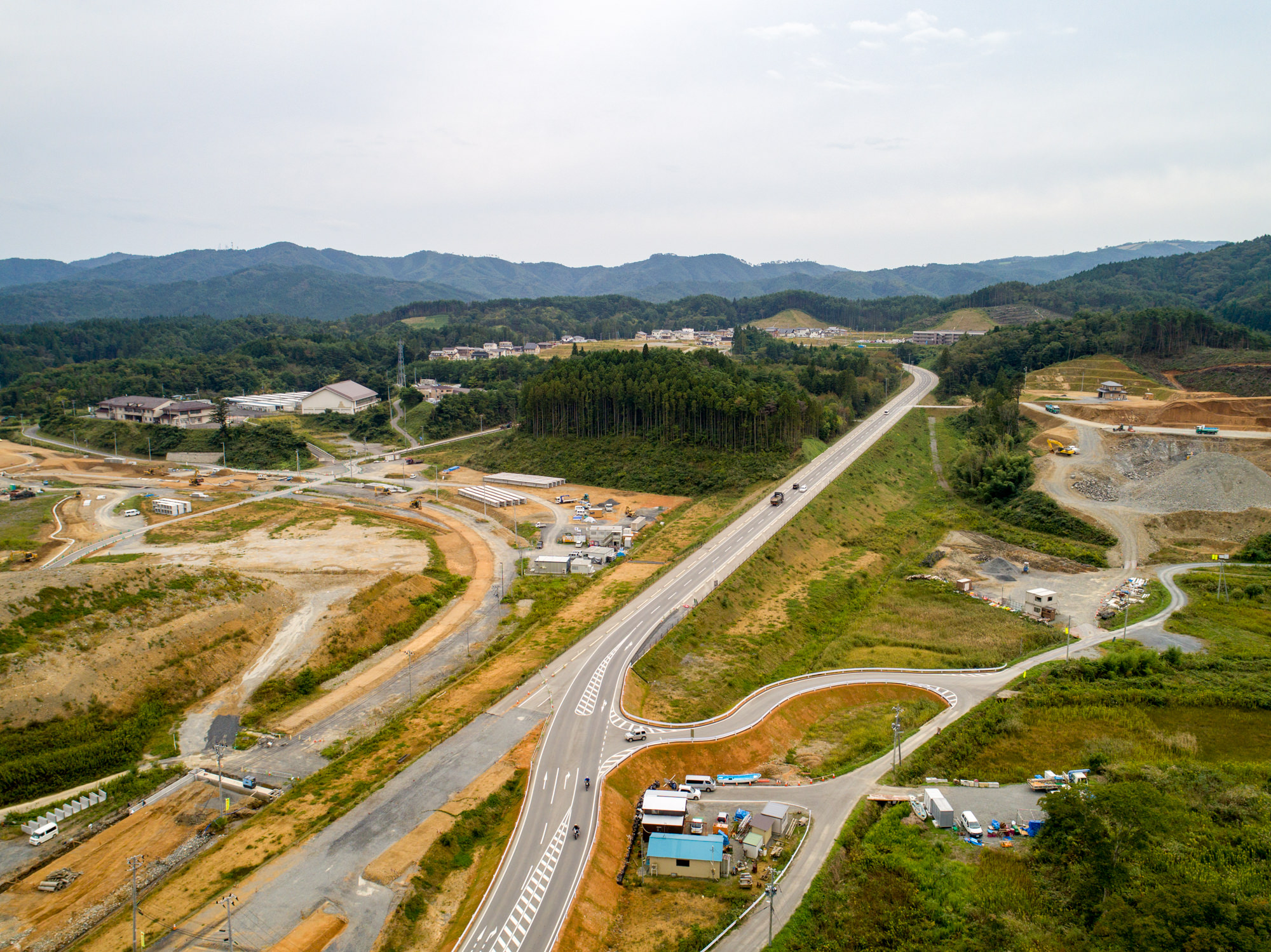 緑に覆われた丘陵地帯を背景に、曲がりくねった道路が田園風景を横切りながら、点在する複数の建設現場や建物、小さな集落へと続く様子を上空から捉えた写真