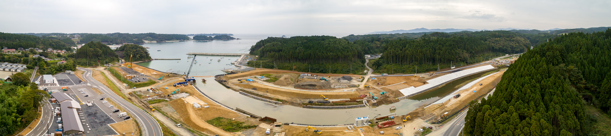 川や運河のような水辺の近くで、クレーンやトラックなどの建設車両が稼働し、道路や水路などのインフラ整備が進められている工事現場を、森林に囲まれた丘陵地と左側に建物や駐車場が広がる風景とともに上空から捉えたパノラマ写真