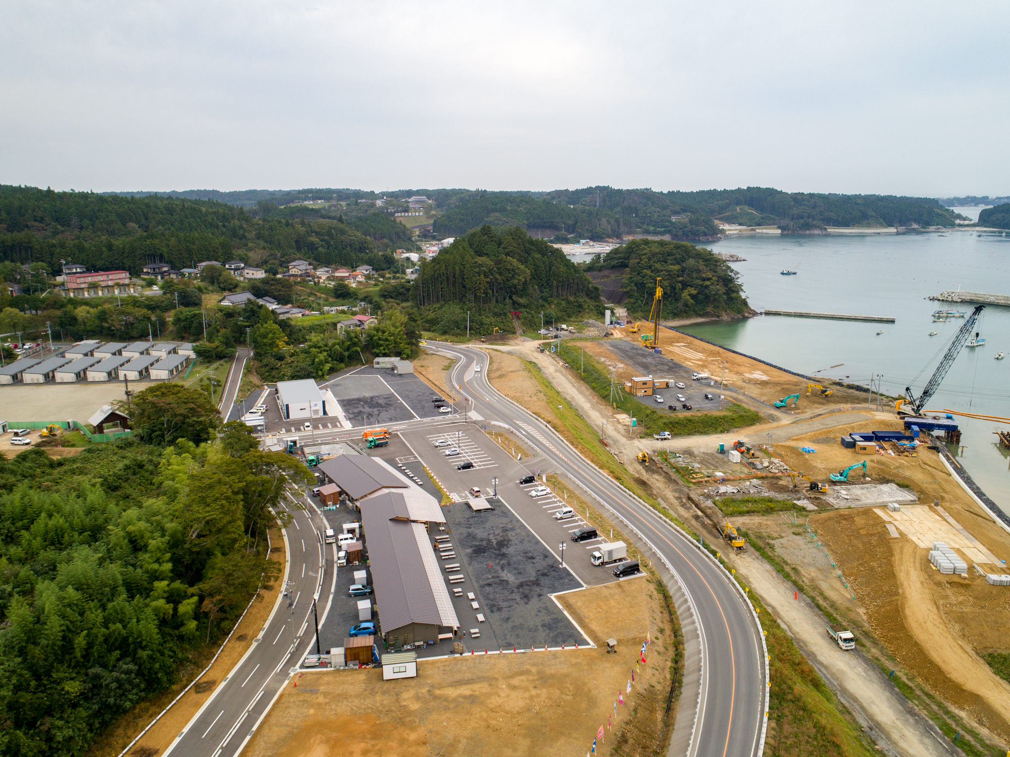 手前に複数の建物や駐車場、道路が広がり、奥には森林や住宅地、海が見え、海岸付近では建設作業が行われている様子を上空から捉えた工事現場の写真