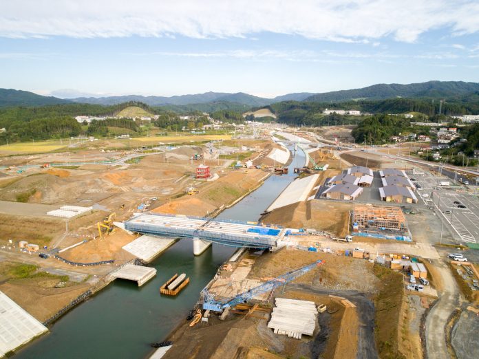 川の上に架かる橋とその周辺で、重機や資材が並び大規模な建設工事が進行している様子を上空から撮影した工事現場の写真