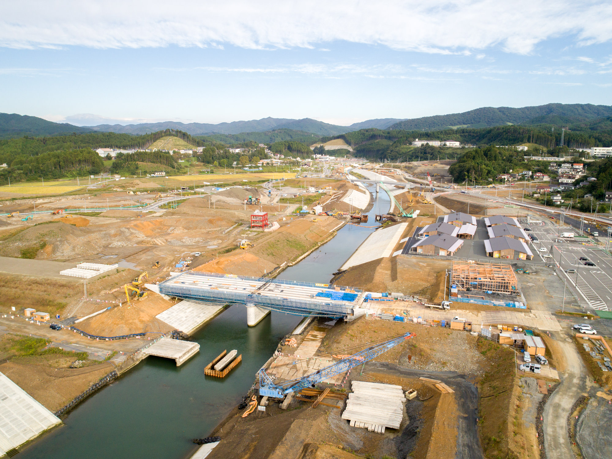 川の上に橋を建設中の現場で、クレーンや足場、コンクリート構造物などの建設資材や機器が点在し、周囲には畑や丘、遠くに建物が見える風景を捉えた工事現場の写真