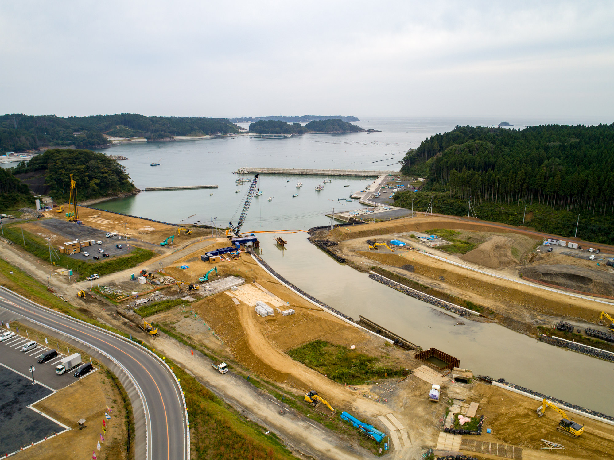 港に複数の船が停泊し、クレーンやショベルカーなどの建設機械が稼働する埋立やインフラ整備中の工事現場を中心に、道路や整地された土地、建設資材が広がり、周囲には森林や遠くに小さな島々が見える工事現場を上空から捉えた写真