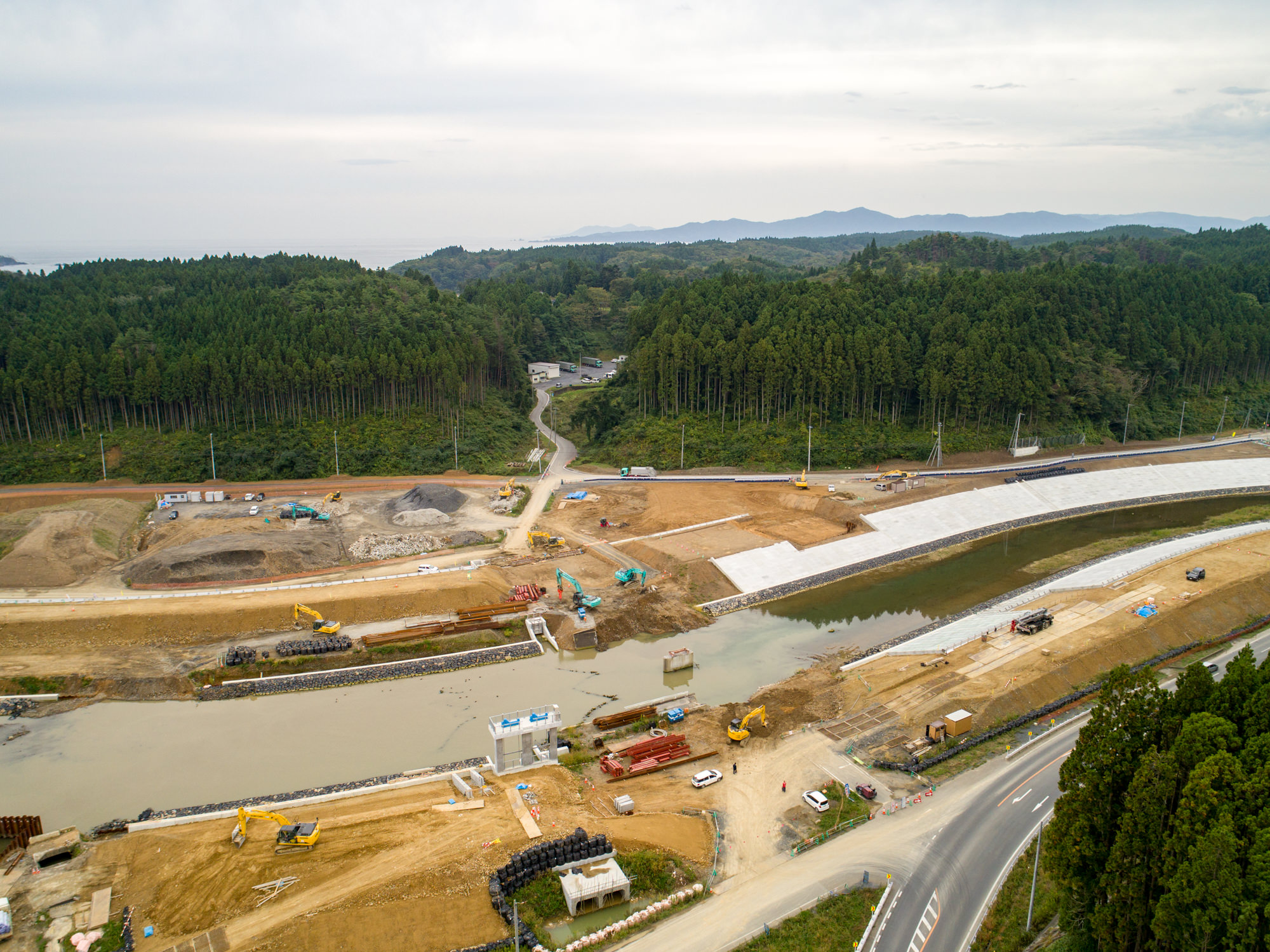 中央に川が流れ、複数の橋や構造物が建設中で、建設機械や設備が点在する工事現場を、周囲の森林と背景の丘陵地とともに上空から捉えた写真