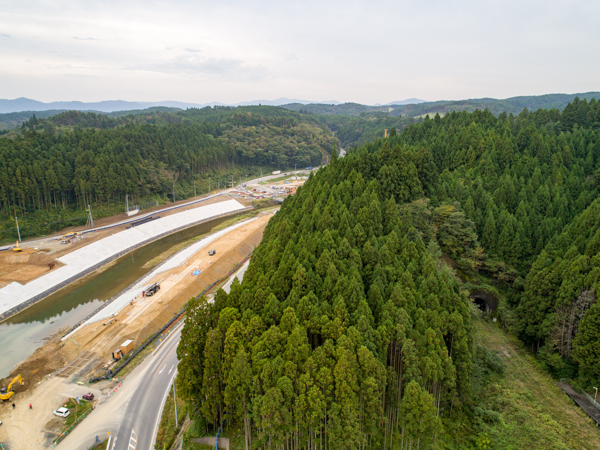 右側に背の高い木々が密集する森林が広がり、左側には道路や重機、川沿いの堤防のような構造物を含む建設現場があり、背景には森林に覆われた丘陵地が広がる風景を曇り空の下で上空から捉えた写真