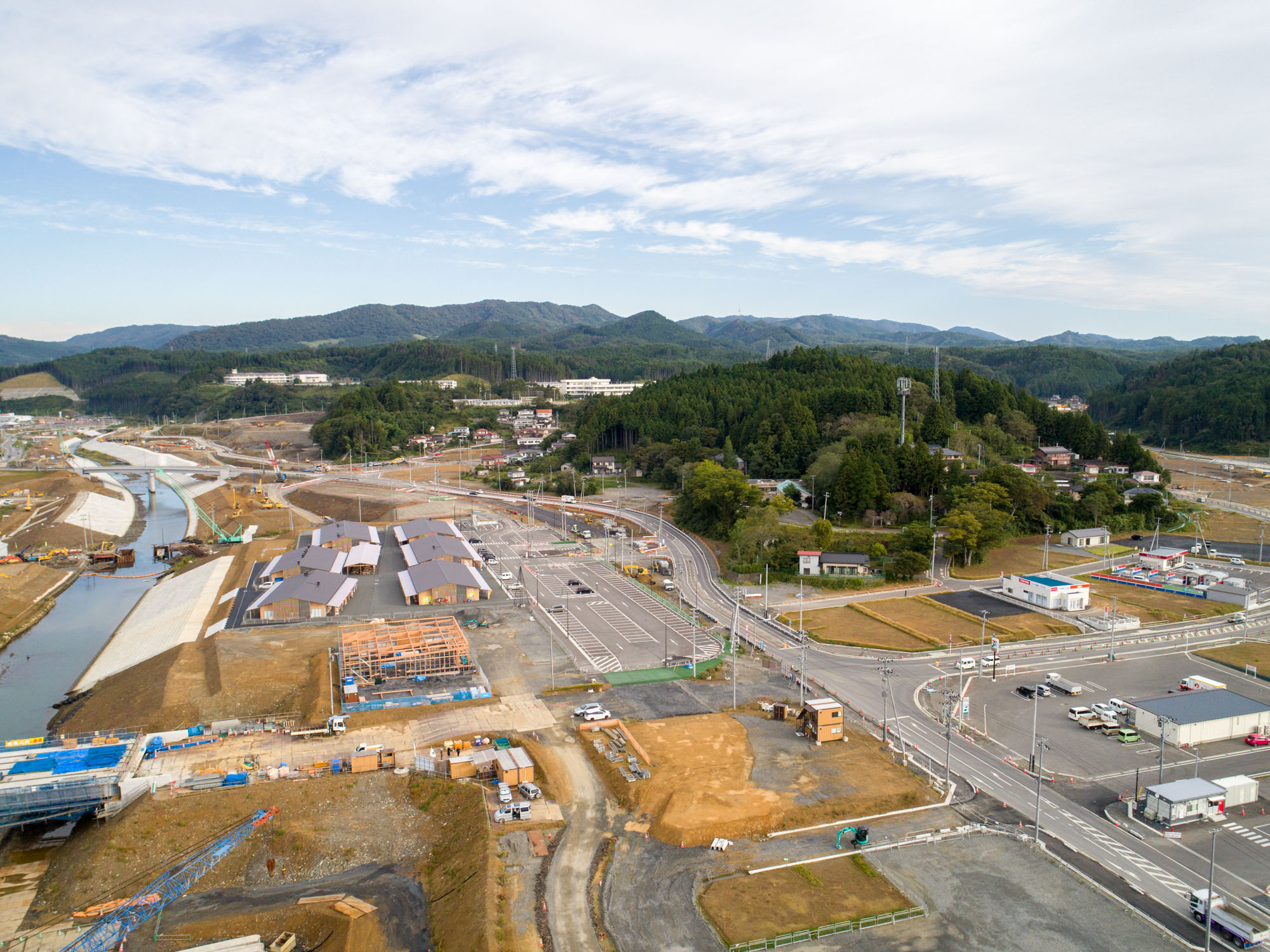 複数の建物が建設中で、道路や川が風景の中を通り、背景には森林に覆われた丘や山々が広がる志津川地区を上空から捉えた工事現場の写真