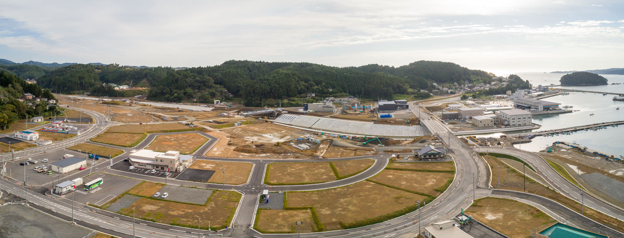 右側に港や海辺が広がり、複数の道路が交差する地域に建物や車両、建設現場、緑地が点在し、背景には木々に覆われた丘が見える沿岸地域を上空から捉えたパノラマ写真